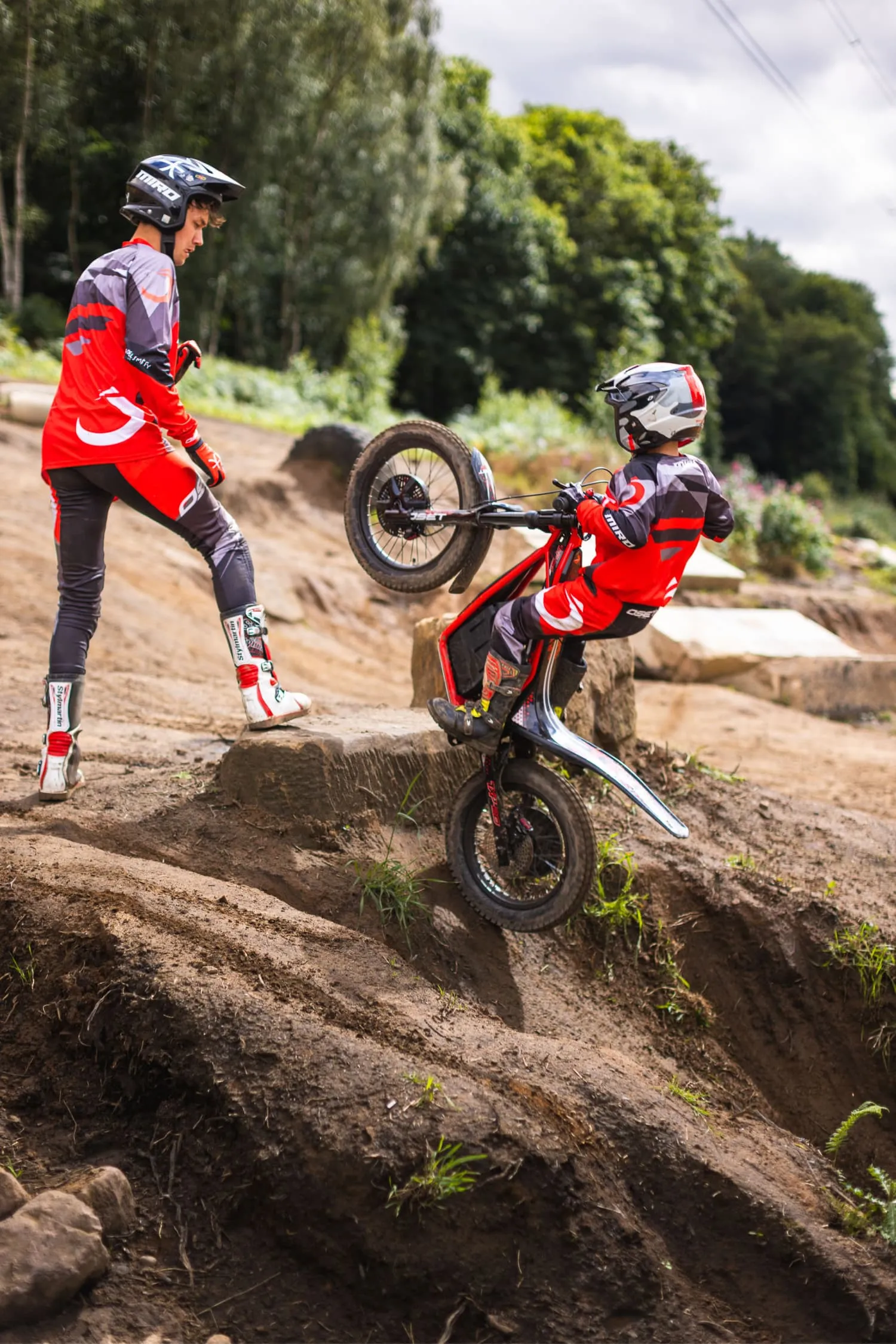 Zwei Motocross-Fahrer in rot-schwarzer Ausrüstung; einer fährt mit angehobenem Vorderrad einen Hang hinauf, der andere steht neben einem Felsen und schaut zu.